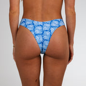 A person is shown from the waist down wearing Honey Swim Coastline | Cheeky Bottoms in blue with a white abstract leaf print, featuring minimal coverage for a sun-kissed look against a plain white background.