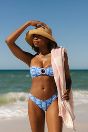 Wearing Honey Swim’s Coastline | Classic Bottoms and a straw hat, a woman stands on a sunny beach with a pink-and-white towel, channeling vintage surf vibes as she looks across the blue ocean and sky.