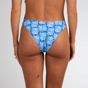 A person faces away, wearing Honey Swim Coastline | Classic Bottoms in blue with a white leaf design. Image shows only their lower back and hips against a plain white background.