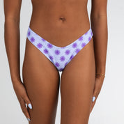 A model poses with hands by their sides in Honey Swims Daybreak | Cheeky Bottoms, featuring a white fabric with a purple sunburst pattern, against a plain white background.