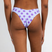 A person faces away, wearing Honey Swims Daybreak | Cheeky Bottoms—a light purple bikini bottom with dark purple palm trees—hands by their sides against a plain white background.
