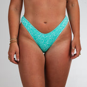 A person models Honey Swims Drift Ribbed | Classic Bottoms in light blue, revealing tan lines on their hips and upper thighs. Their ring- and bracelet-adorned hands rest at their sides against a plain white backdrop.