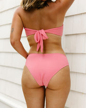 A person faces away outdoors wearing the Honey Swim Flamingo Ribbed Bandeau Set with a back bow tie, arm raised to show a small left upper arm tattoo; white siding is in the background. badge text: { Wearing Medium Top & Full Coverage Medium Bottoms}