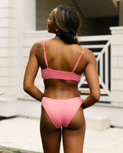A woman stands outdoors with her back to the camera, wearing the Honey Swim Flamingo Ribbed Bralette Set. She has long hair, a small left shoulder tattoo, and light skin patches. A white building is visible behind her.