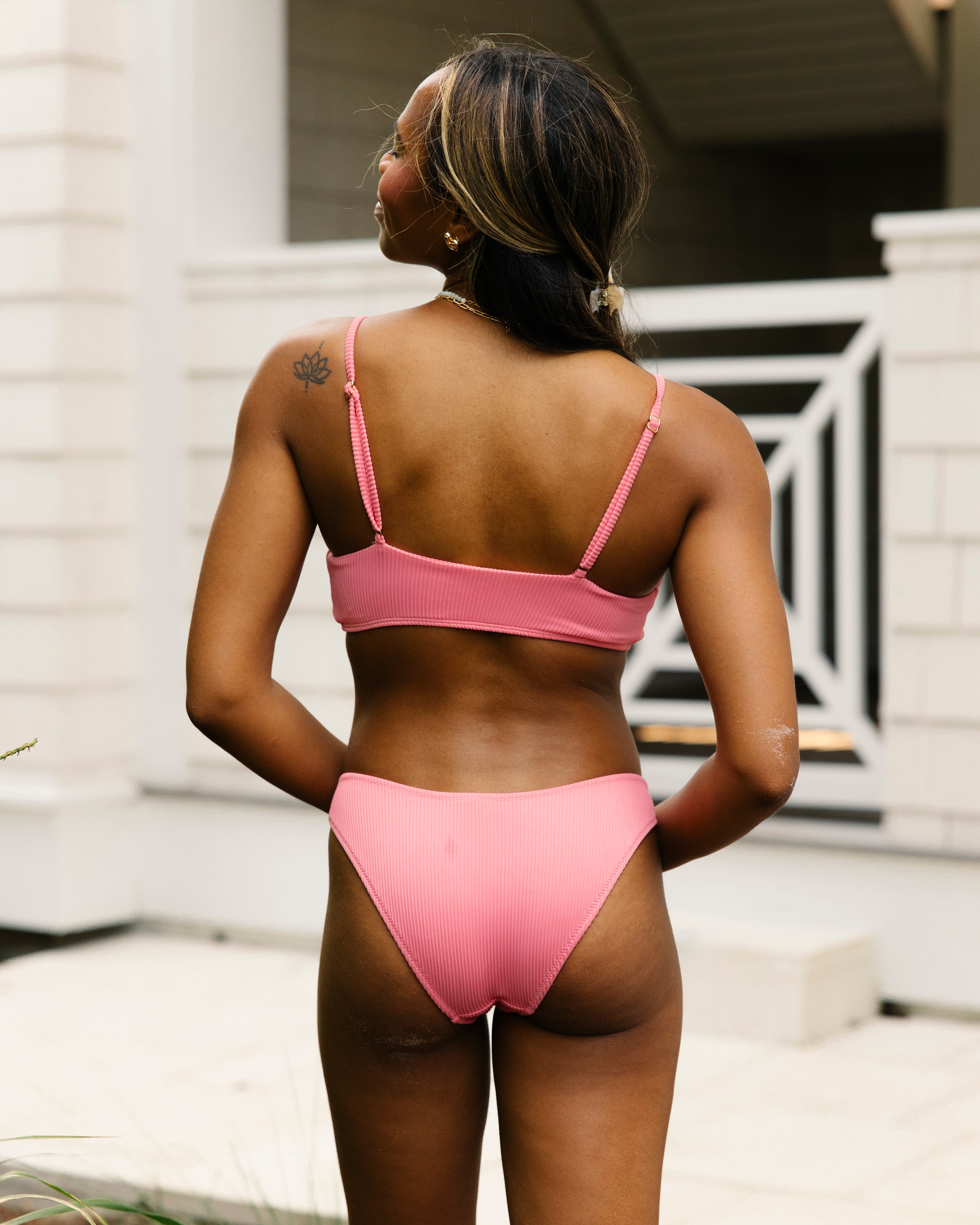 A woman stands outdoors with her back to the camera, wearing the Honey Swim Flamingo Ribbed Bralette Set. She has long hair, a small left shoulder tattoo, and light skin patches. A white building is visible behind her.