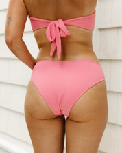 A person models the Honey Swim Flamingo Ribbed Bottoms, back to the camera, highlighting the bow-tied top and ribbed texture near a light-paneled wall.