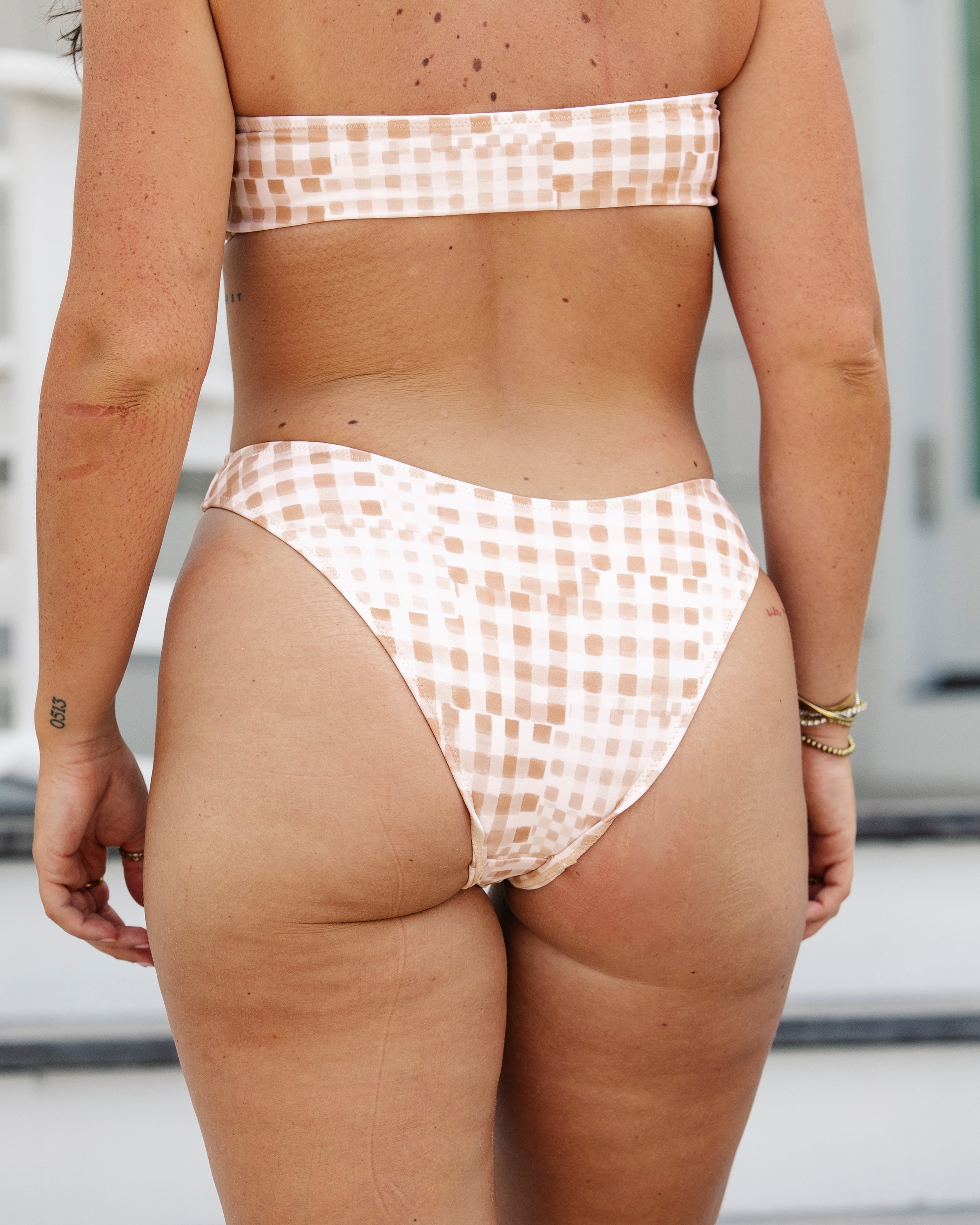 Outdoors, a woman models Honey Swim’s Harper | Bottoms – All Styles, featuring a light geometric grid pattern and high-waisted fit; the photo highlights her back, legs, and natural skin texture.