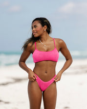 A woman wears the Honey Swim Hot Pink Sporty Luxe Top on a sandy beach, glancing to the side with a slight smile as the ocean and sky create a stunning backdrop.