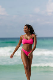 A woman in the Hot Pink Sporty Luxe Top by Honey Swim smiles as she walks along the beach, with ocean waves and a partly cloudy sky behind her.