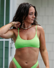A woman with long dark hair models the Honey Swim Kiwi Ribbed Bralette Set, layered with jewelry, standing outdoors before a light-colored building and looking to her left.