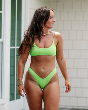 A woman with long dark hair wears the Honey Swim Kiwi Ribbed Bralette Set, accessorized with bracelets, necklaces, and earrings. She stands outdoors by a white building with large windows, looking to her right.