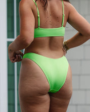A woman wearing Honey Swim Kiwi Ribbed Bottoms stands outdoors with her back to the camera near a white building and glass window, showing her arms, back, and legs.