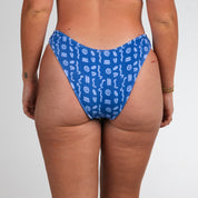 A person stands facing away, wearing Honey Swims Lagoon Ribbed | Classic Bottoms. The ribbed texture adds interest, while small tattoos and a gold bracelet accent the relaxed pose against a plain white background.