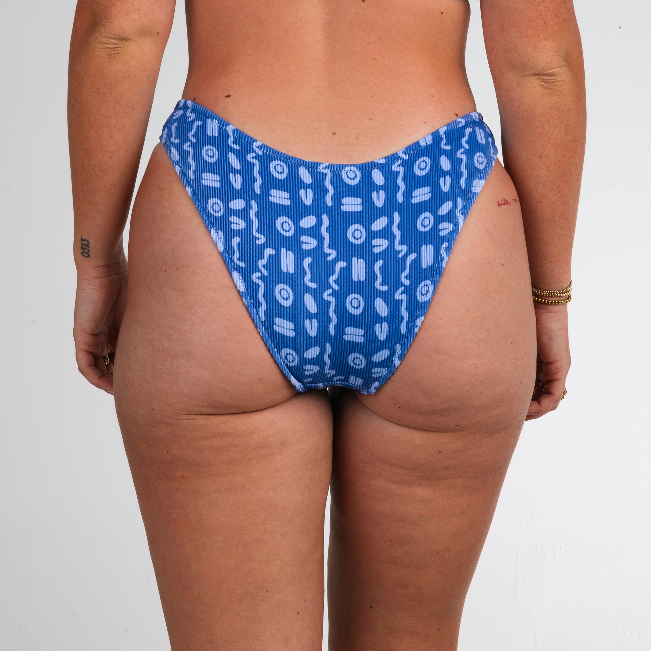 A person stands facing away, wearing Honey Swims Lagoon Ribbed | Classic Bottoms. The ribbed texture adds interest, while small tattoos and a gold bracelet accent the relaxed pose against a plain white background.