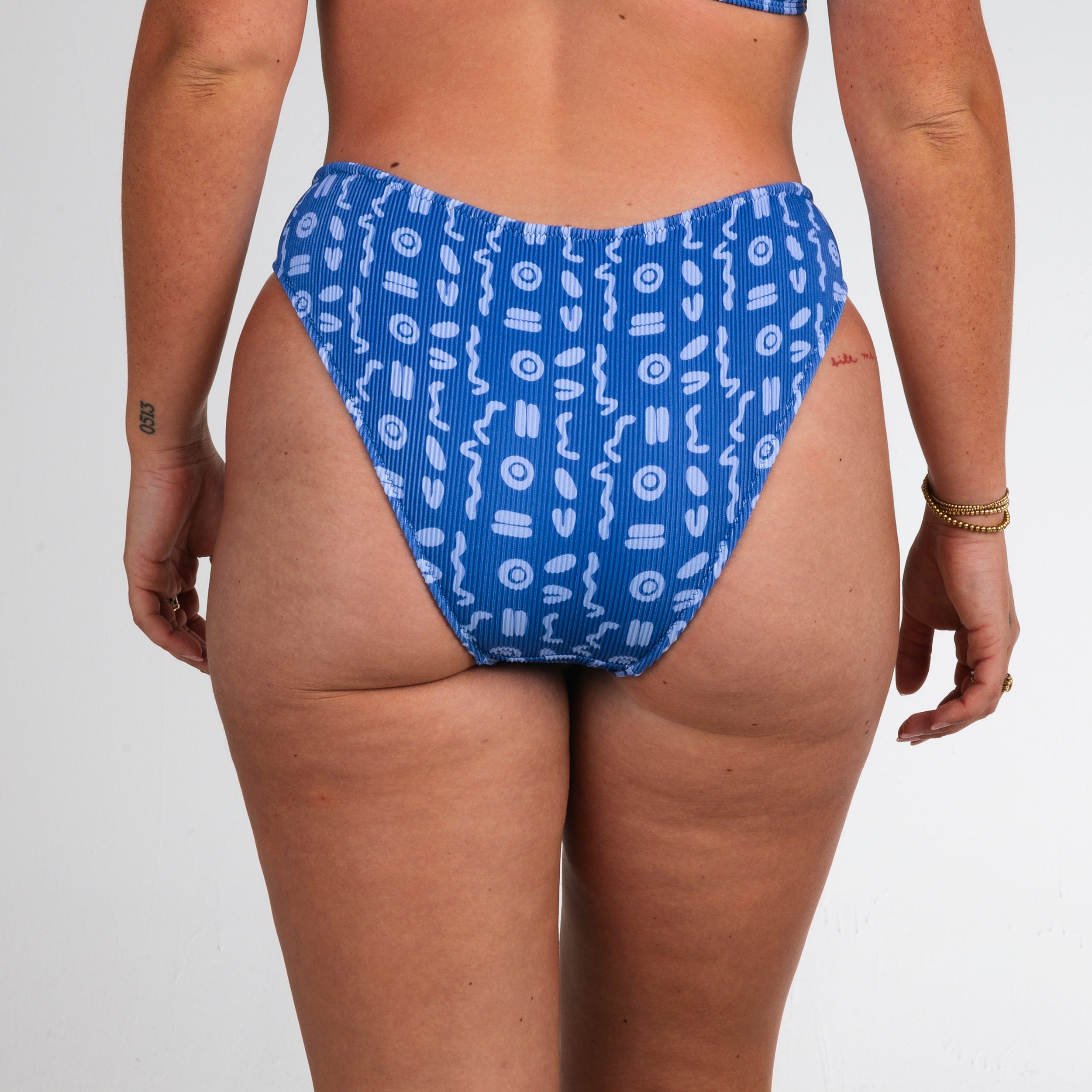 A person models the Honey Swim Lagoon Ribbed | Full Coverage Bottoms, featuring white geometric patterns, facing away to show the lower back and legs against a plain light background.