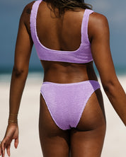 A woman stands on a sandy beach facing away, wearing the Honey Swim Lilac Luxe | Classic Bottom. The background shows a blurred ocean and sky, and her hair falls loosely.