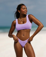 A woman smiles with her eyes closed, facing the sun on a sandy beach, wearing the Lilac Luxe | Top by Honey Swim. The ocean and blue sky create a blurred background.