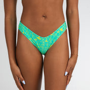 A model wears Honey Swims Lush | Cheeky Bottoms in turquoise with a yellow tropical print, standing against a plain backdrop, showing sun-kissed skin, light blue nails, and a delicate bracelet.