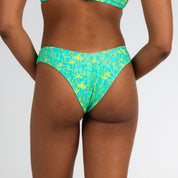A person with sun-tanned skin stands against a plain white background, shown from the back wearing Honey Swim’s Lush | Cheeky Bottoms in green with a yellow abstract pattern.