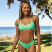 A woman in a green patterned bikini and Honey Swim Lush | Classic Bottoms smiles outdoors near the beach, with palm trees and the ocean behind her on a sunny day.