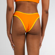 A person faces away, wearing Honey Swims Marigold Terry | Cheeky Bottoms with a matching top. Their nails are light blue, gold bracelets on their wrists, set against a plain white background.