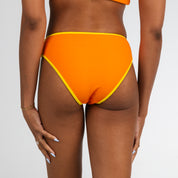 A person with medium brown skin models Honey Swims Marigold Terry Full Coverage Bottoms, a high-waisted orange bikini with yellow trim, shown from the back against a plain white background.
