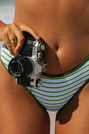 A person wears Honey Swims Piper Ribbed Full Coverage Bottoms in green and white, holding a vintage film camera by their waist. Only their midsection and hand are visible.