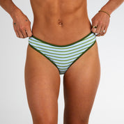 A person models the Honey Swim Piper Ribbed Full Coverage Bottoms in green and white stripes, standing against a white background with rings and a small tattoo visible on their left hand.