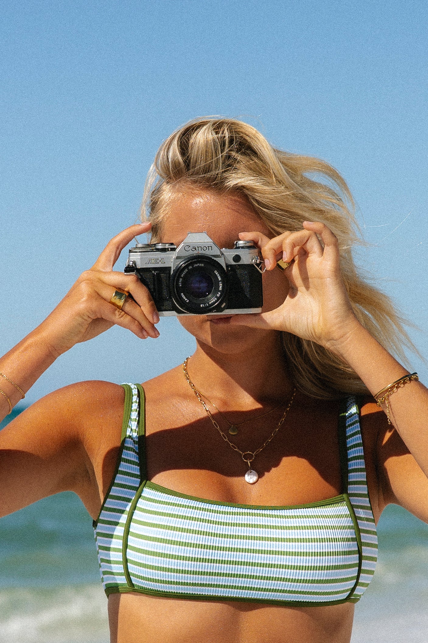 A woman wearing the Honey Swim Piper Ribbed Squareneck Top holds a vintage Canon camera to her face, capturing moments on a tropical beach with blue skies and ocean behind her.