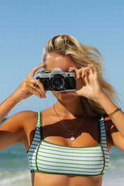 A woman wearing the Honey Swim Piper Ribbed Squareneck Top holds a vintage Canon camera to her face, capturing moments on a tropical beach with blue skies and ocean behind her.