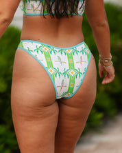 A woman models the Honey Swim Polly Palm Bottoms, a high-waisted bikini featuring palm tree and parrot motifs, standing outdoors among lush greenery with her face out of frame.