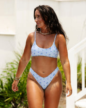 A woman with long dark hair wears the Posie Bralette Set by Honey Swim, posing outdoors near white stairs with greenery and a light-colored building in the background.