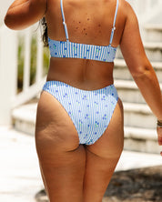 A woman wears the Posie bikini bottoms by Honey Swim in blue and white stripes with floral patterns, walking outdoors near white stairs in bright sunlight, seen from the back.