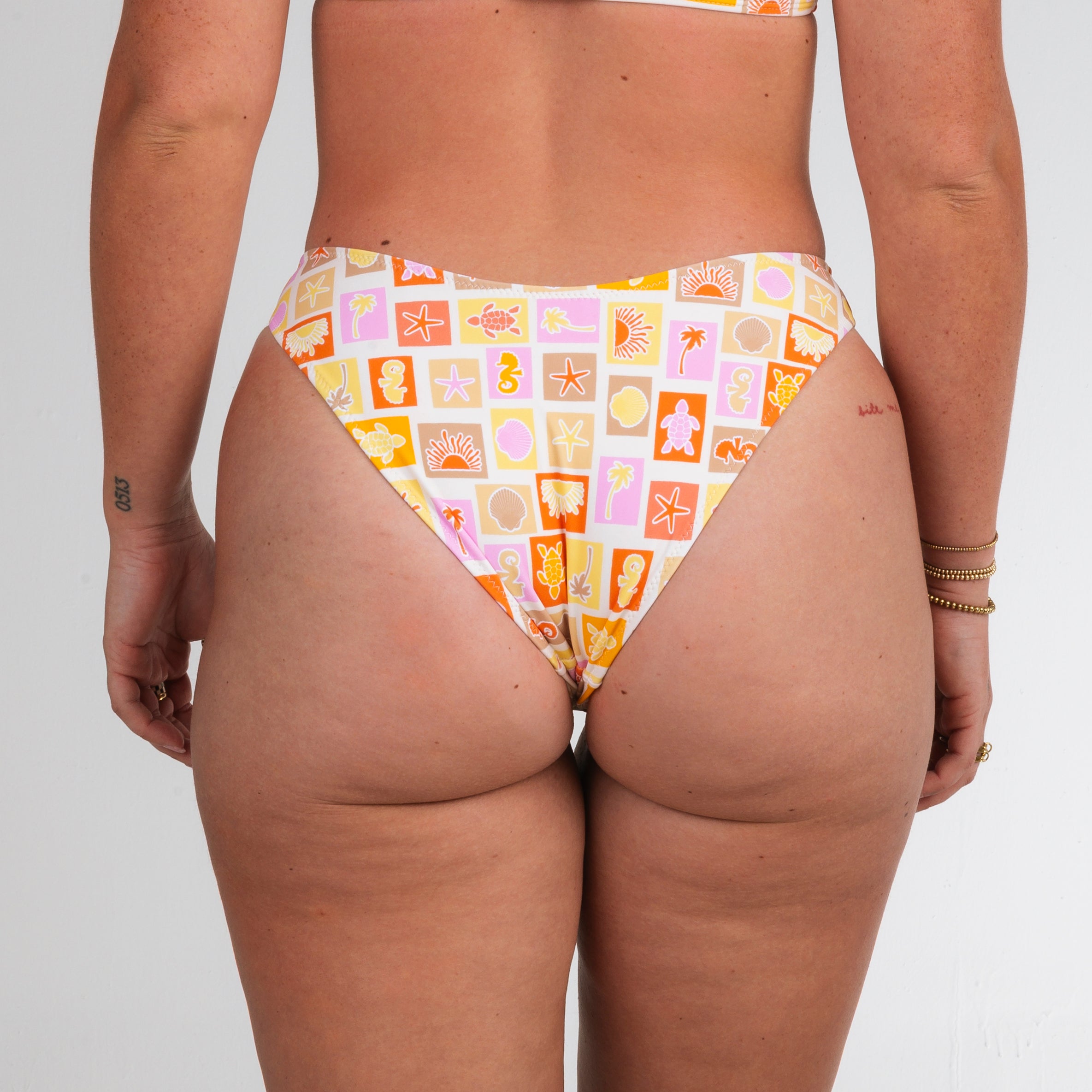 Someone models the Honey Swim Resort | Cheeky Bottoms with colorful geometric and abstract prints, photographed from the back against a white background for a true beach vibe.