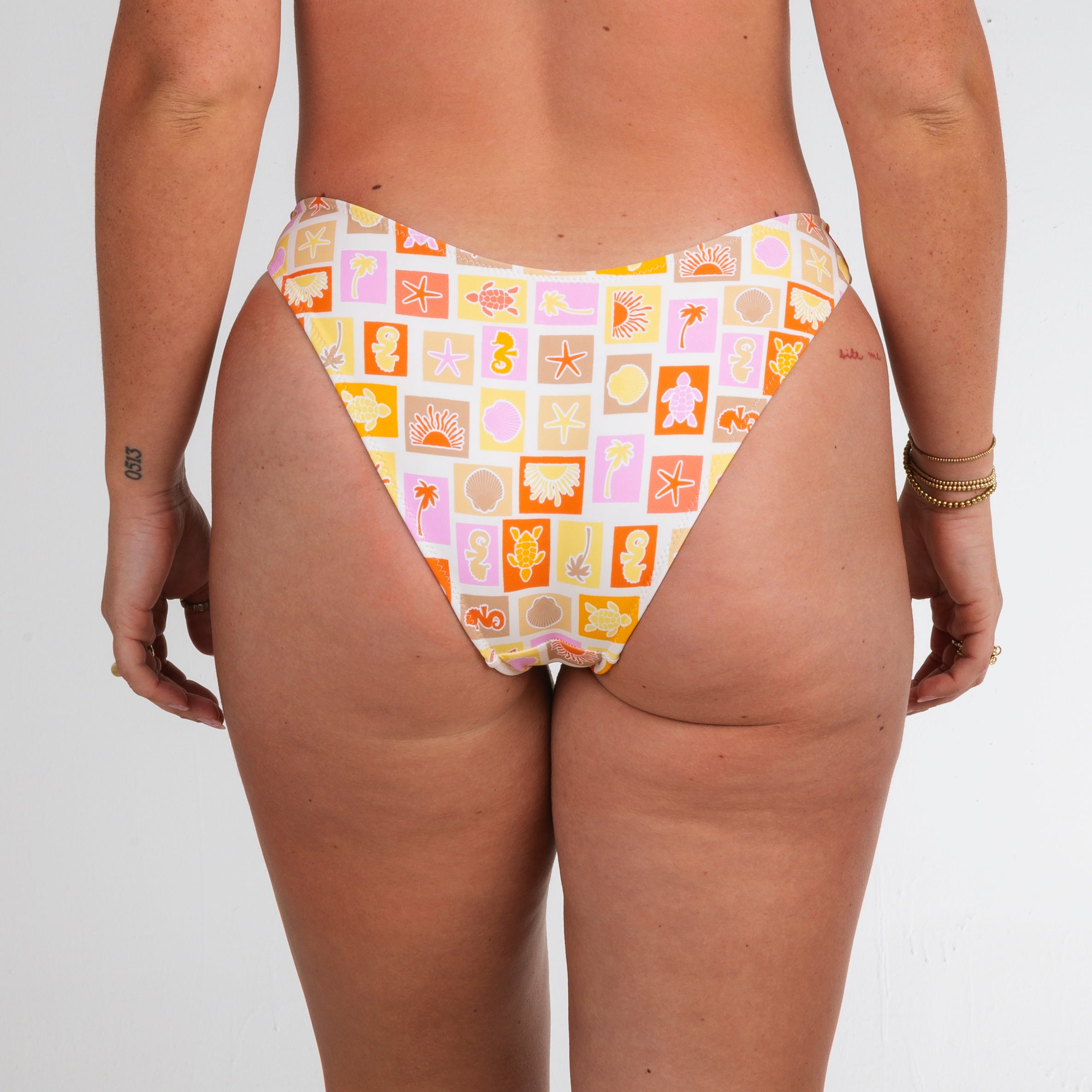 A person models Honey Swim’s Resort | Classic Bottoms featuring a vibrant sea creature and shell print, shown from the waist down and back view, radiating beach vibes on a plain white background.