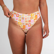 Wearing Honey Swims Resort Highwaisted Bottoms, featuring a colorful, retro tile print of shells, flowers, and abstract shapes, she channels dreamy beach vibes with hands on hips.