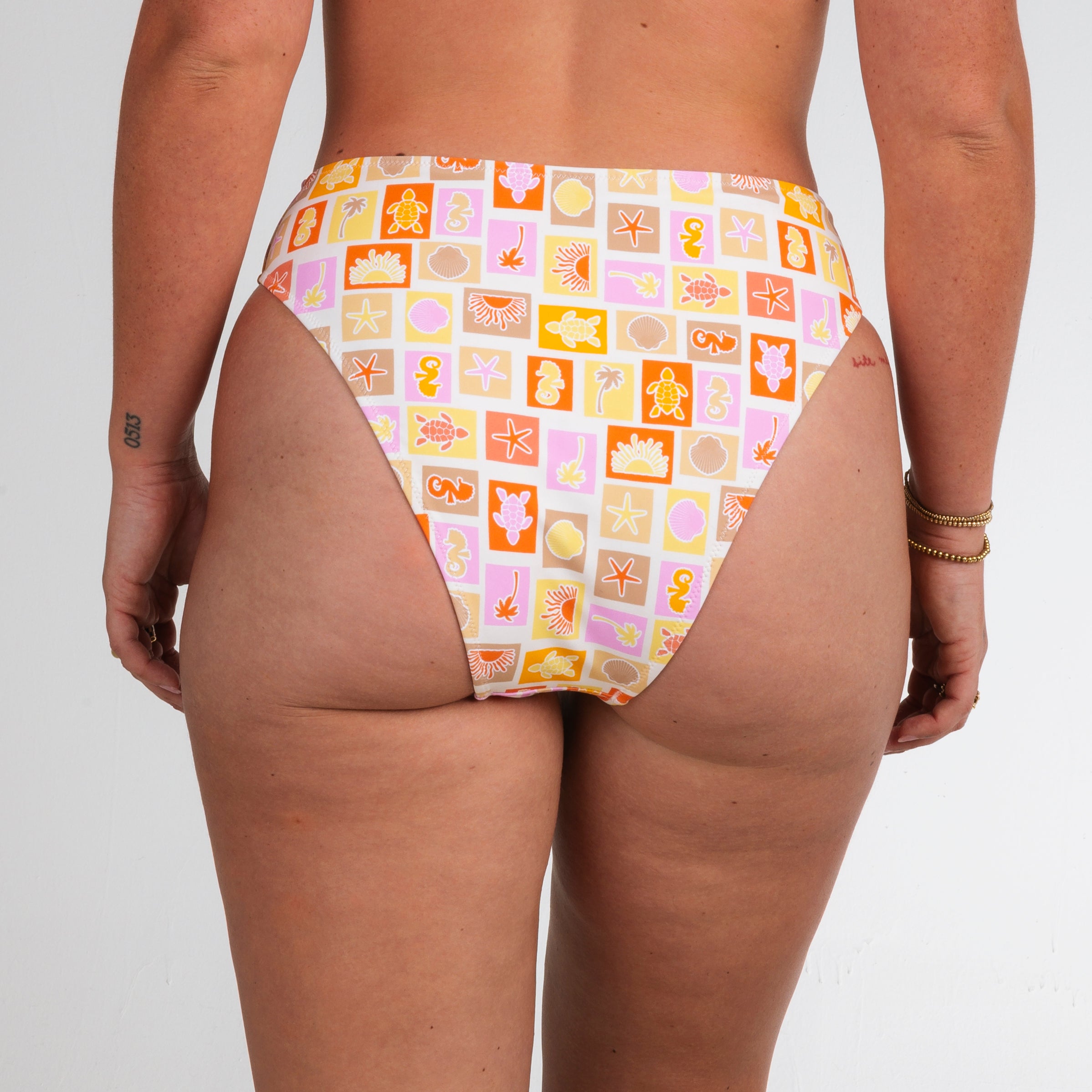 Wearing Honey Swims Resort Highwaisted Bottoms, featuring a colorful geometric print with abstract beach icons in orange, yellow, pink, and white—exuding dreamy beach vibes. Photo from the back.