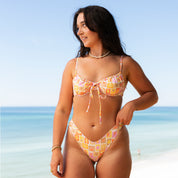 A woman with long brown hair wears the Honey Swim Resort String Underwire Set on the beach, gazing to the side against an ocean and blue sky backdrop.