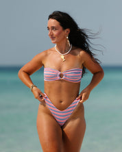 A woman wearing the Sorbet Ribbed Bandeau Set by Honey Swim stands on a beach with clear blue water, her long dark hair flowing as she looks to the side with a relaxed expression.
