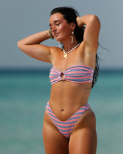 A woman models the Sorbet Ribbed Bandeau Set by Honey Swim on a sunny beach, wearing jewelry and posing with her hands behind her head against an ocean and blue sky backdrop.