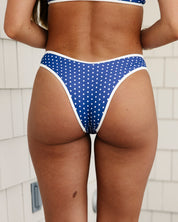 A person faces away from the camera in a cheeky Spritz bikini bottom with white trim by Honey Swim, set against white shiplap-style siding.