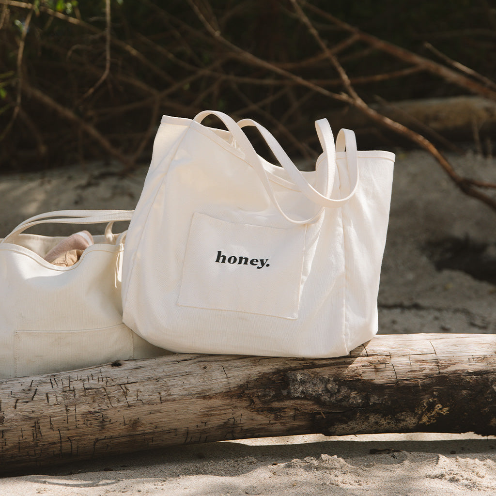 Two white canvas bags are on a driftwood log in a sandy outdoor setting. One is the oversized Honey Tote Bag by Honey Swim, featuring the word honey. on its front pocket, with sparse greenery in the background.