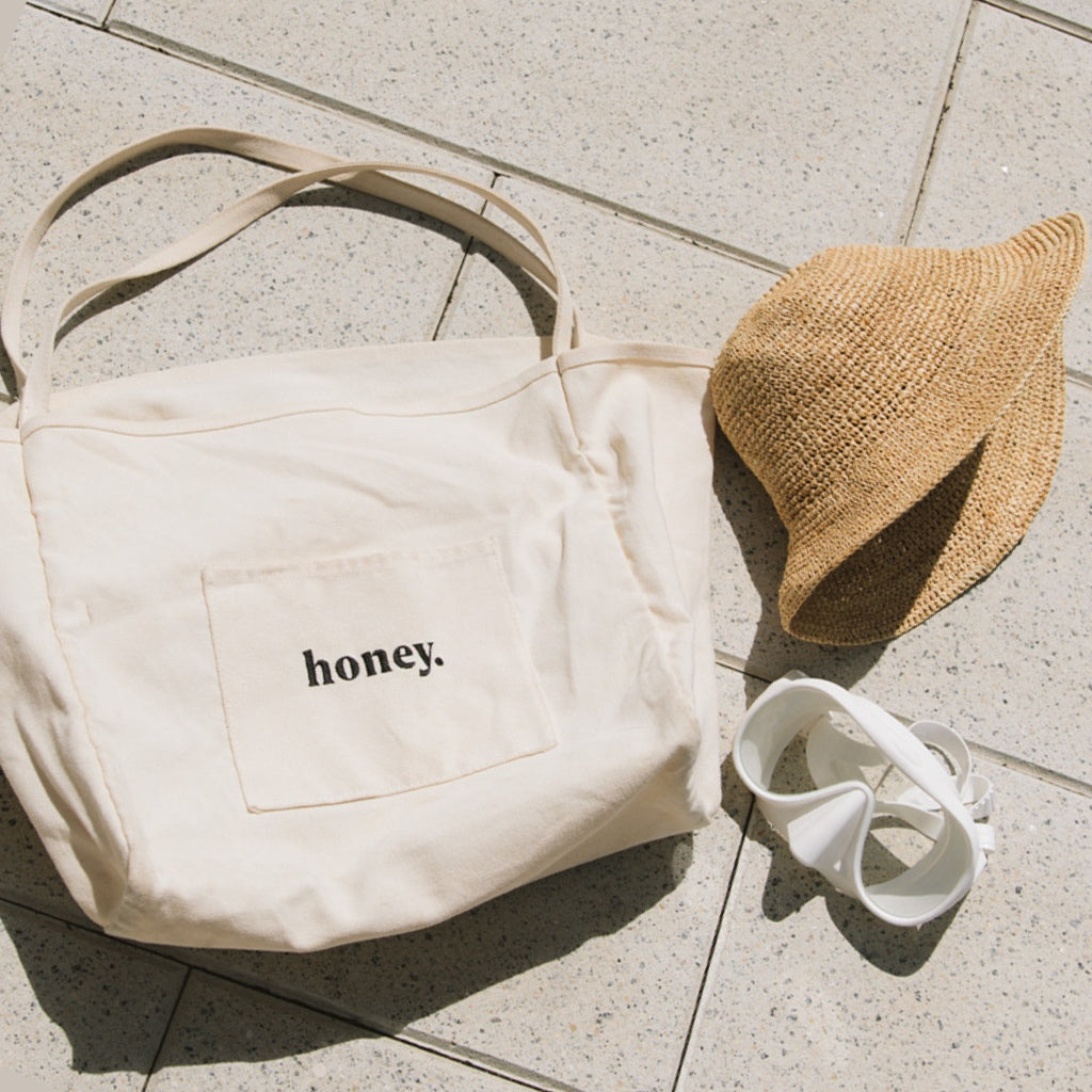 The Beach Essentials Bundle by Honey Swim includes a beige honey. pocket tote, Inlet Hat, and white sunglasses on a tiled surface – perfect for your beach day.