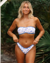 A blonde woman stands outdoors before greenery, wearing Honey Swims Vista Cinched Bandeau Set in a white and blue pattern, accessorized with gold bangles as she adjusts her bikini bottoms.