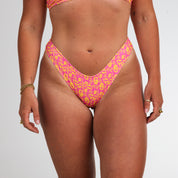 A person models Honey Swims Wildflower | Cheeky Bottoms and a pink and yellow patterned bikini top against a white background, capturing a carefree summer vibe with arms relaxed at their sides.