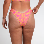 Model wears Honey Swims Wildflower | Cheeky Bottoms, featuring a pink and yellow floral print, shown from the back against a white background with small tattoos and bracelets for a carefree summer look.