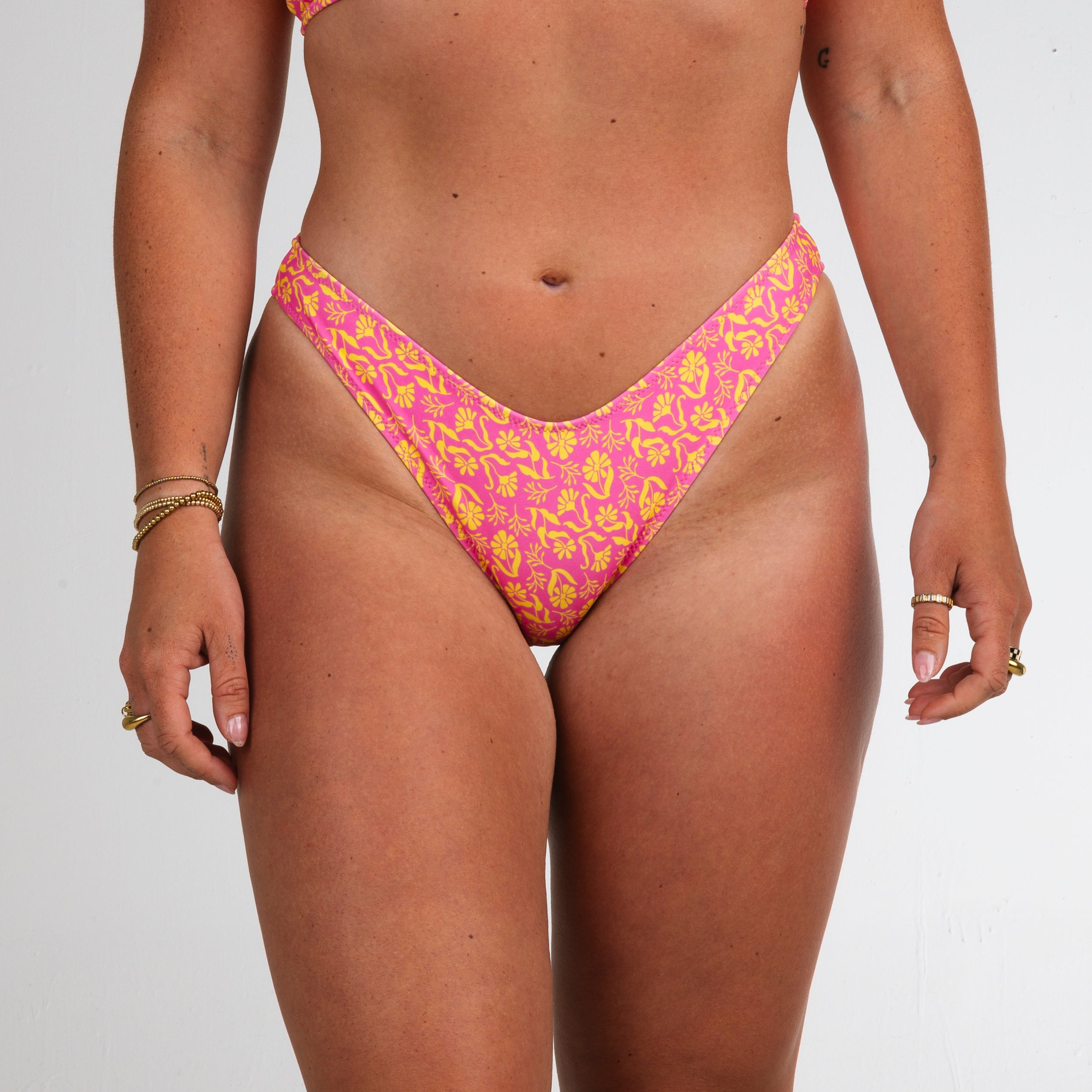A person models Honey Swim’s Wildflower | Classic Bottoms in a pink and yellow pattern, featuring a pop of pink and visible tan lines, standing against a plain white background.