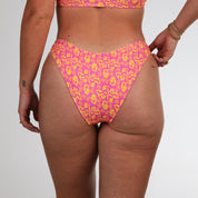 A person wears Honey Swims Wildflower | Classic Bottoms bikini, featuring pink and yellow hues. Their hand with gold bracelets rests on their hip, set against a plain white background.