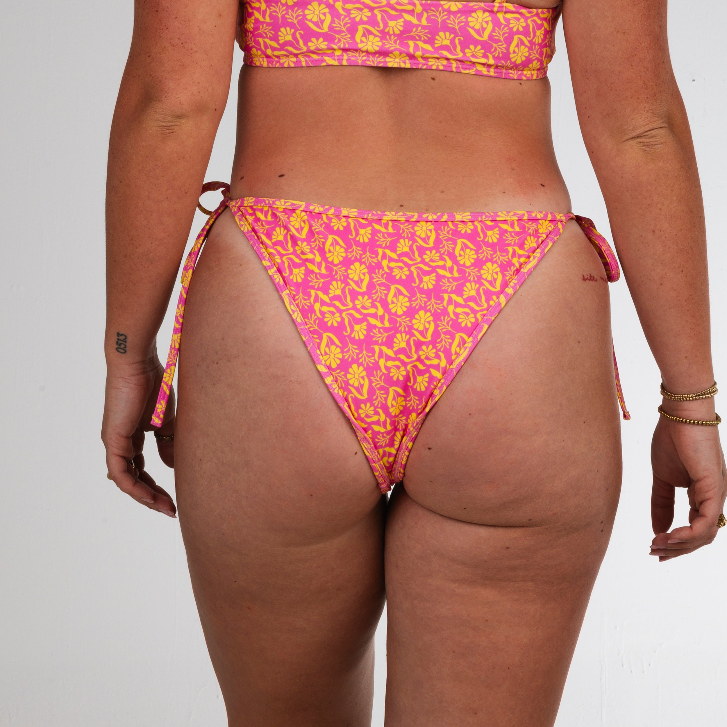 A person faces away from the camera, wearing Honey Swim’s Wildflower | String Bottoms in pink and yellow floral print with adjustable side ties, arms relaxed by their sides, against a plain white backdrop.