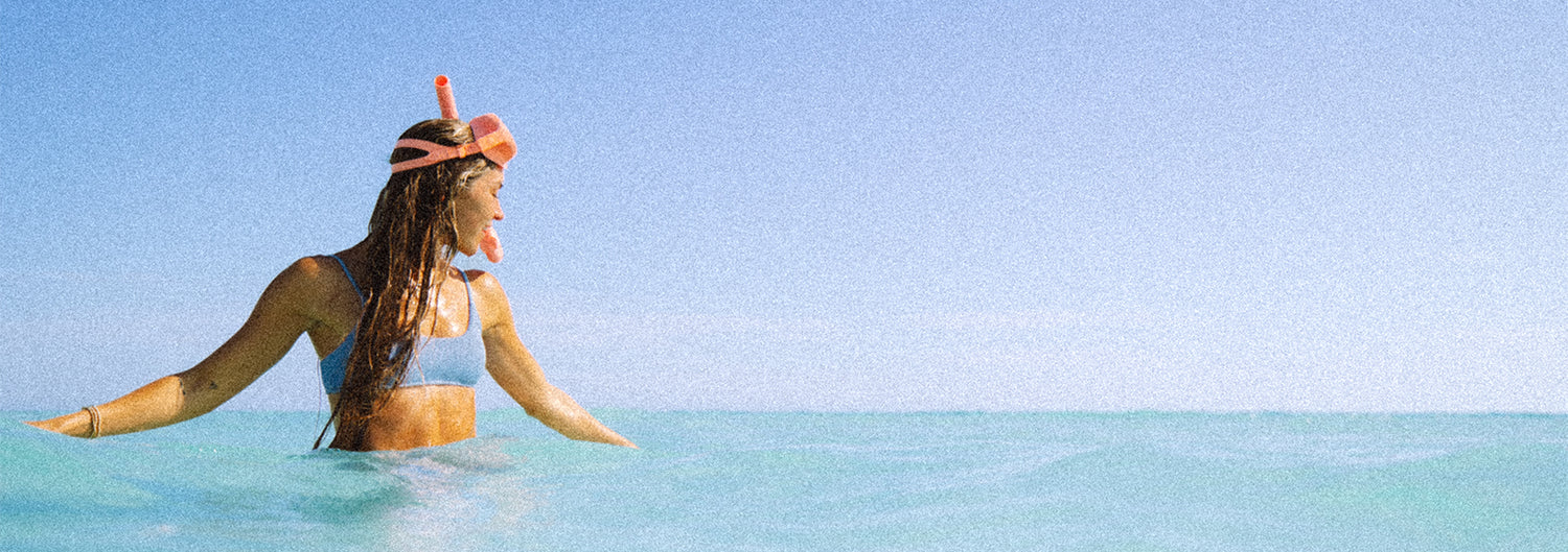 A person stands in clear, turquoise ocean water wearing a snorkel mask and a swimsuit. The sky is clear and blue, creating a serene atmosphere.